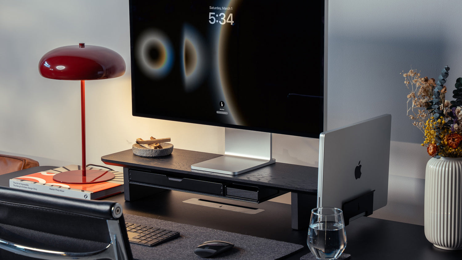 Totalaufnahme des Setup Cockpit Monitorständers Medium in All Black. Ausgestattet mit Laptop Dock, Großsem Fach und Ablage. In modernem Office Setting mit Desk Pad, Vitra Schreibtischstuhl und roter Tischleuchte.