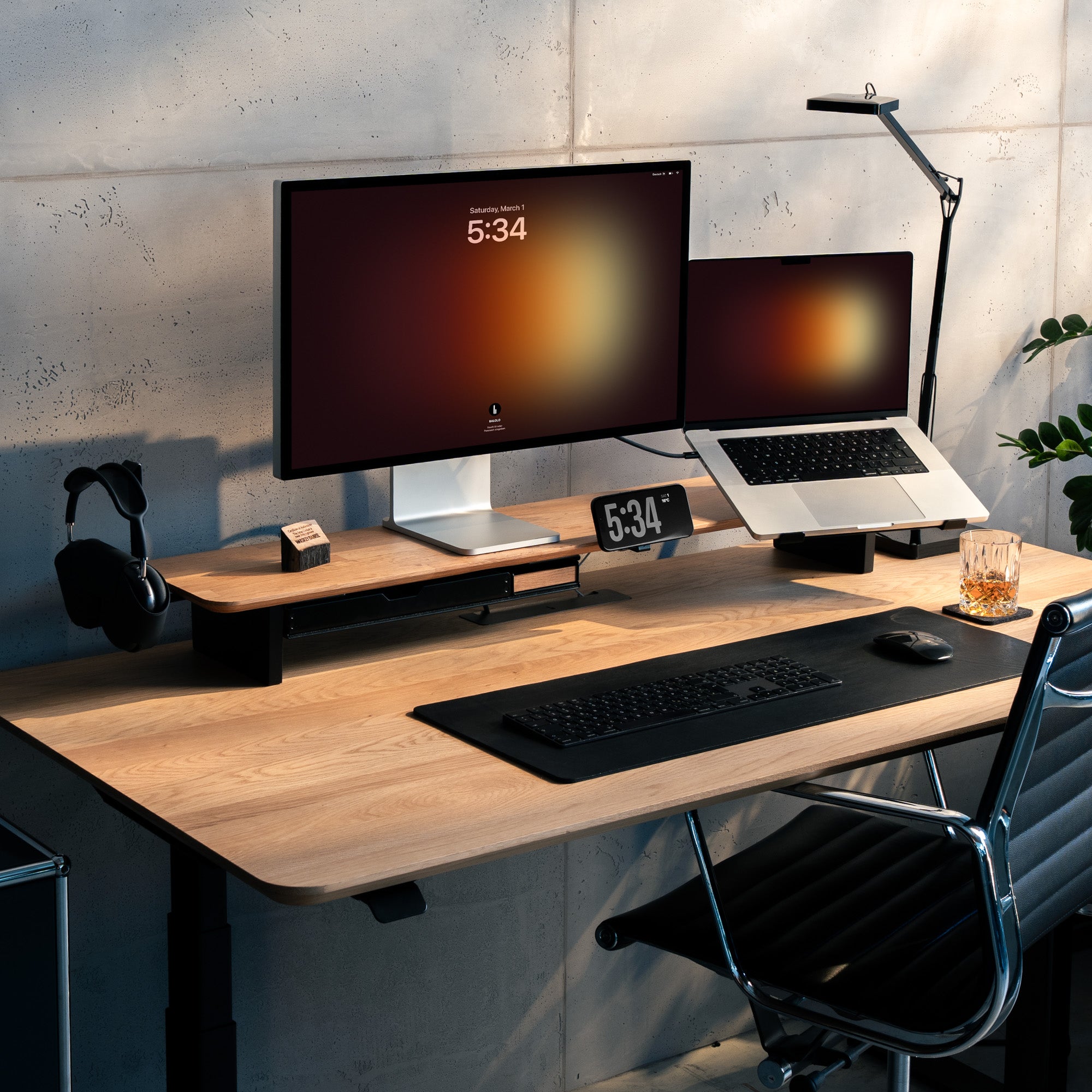 Beauty Shot des Setup Cockpit Monitorständers als Whiskey Barrel Edition. Inszeniert im modernen Home Office Setting mit Apple Studio Display und Macbook. Mit Anbauteilen wie Kopfhörerhalter, Fach und Handyhalterung.