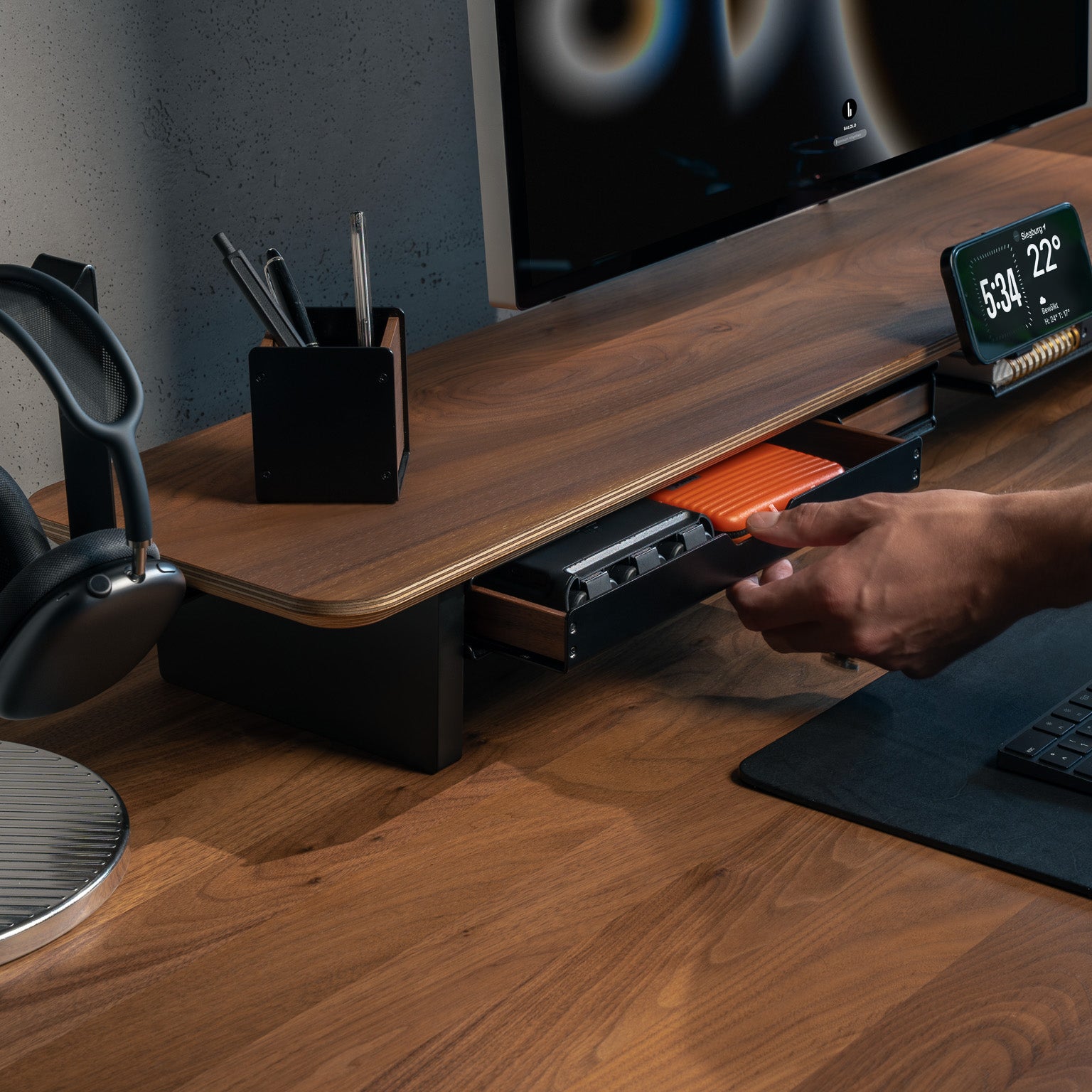 Detailaufnahme des Setup Cockpit Monitorständers, Person zieht große Stiftablage aus montiertem Fach. Ergänzt durch Stifthalter, Kopfhörerhalter, MagSafe Halter und Apple Studio Display in modernem Office Setting.